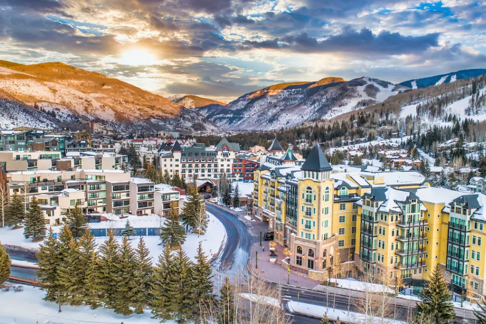 Aerial view of Vail, Colorado ski resort town with multi-story buildings in foreground and snowy mountains in background.
